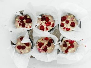 Muffin tray with six equal portions of muffin mix topped with raspberries and white chocolate bits