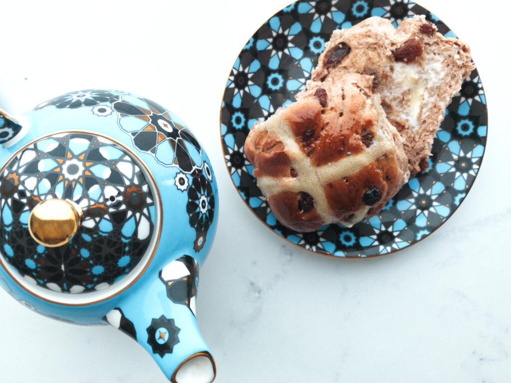 Hot cross bun on plate with lashing of butter sitting next to a blue teapot